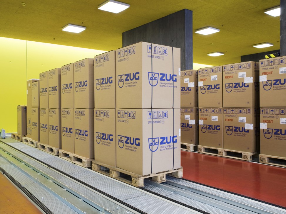 Stacked cardboard boxes labeled V-ZUG with 'SWISS MADE' marking in a warehouse with yellow walls.