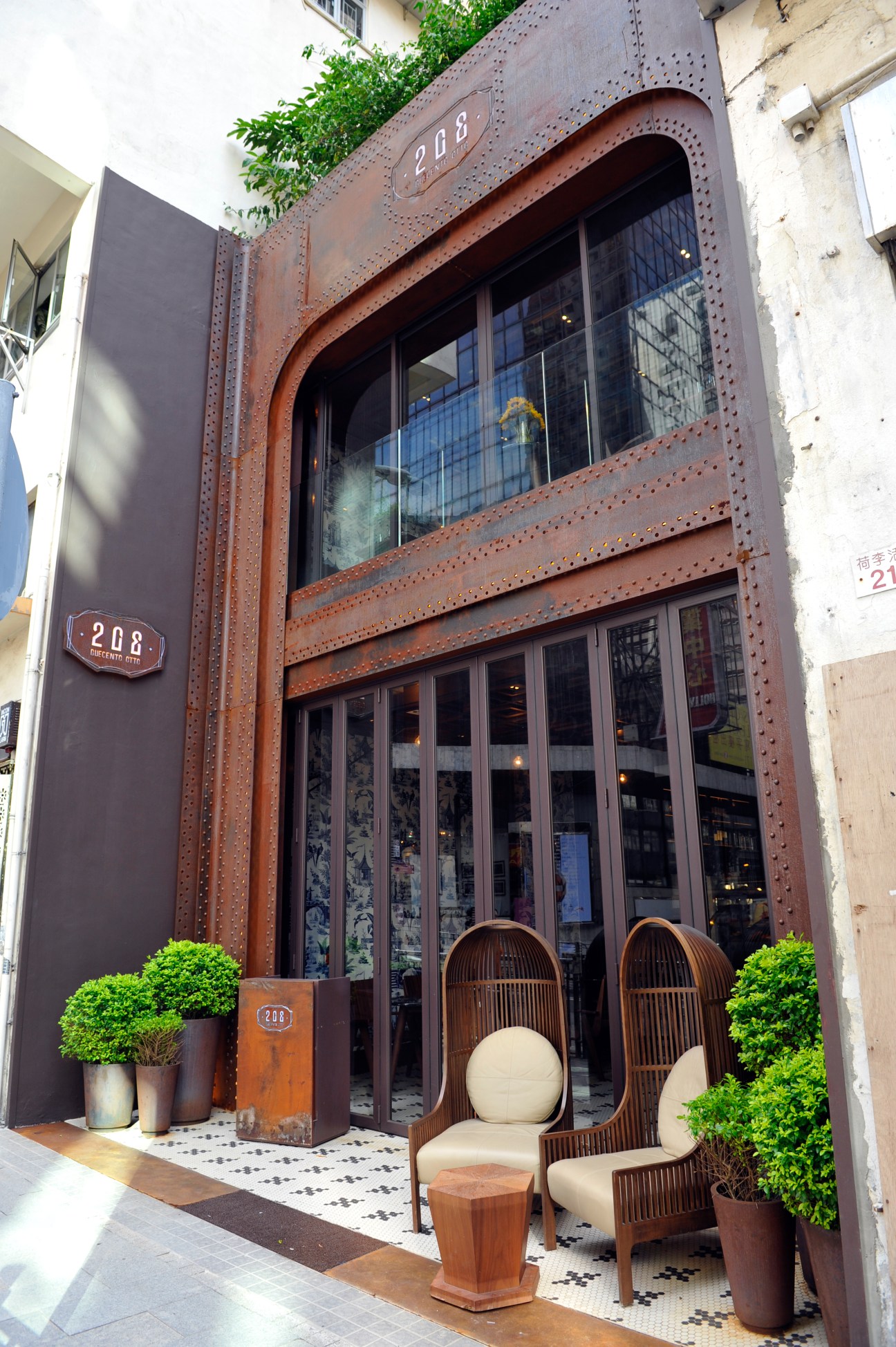 Storefront of 208 Duecento Otto restaurant with rusted metal facade, large windows, and wooden chairs outside.
