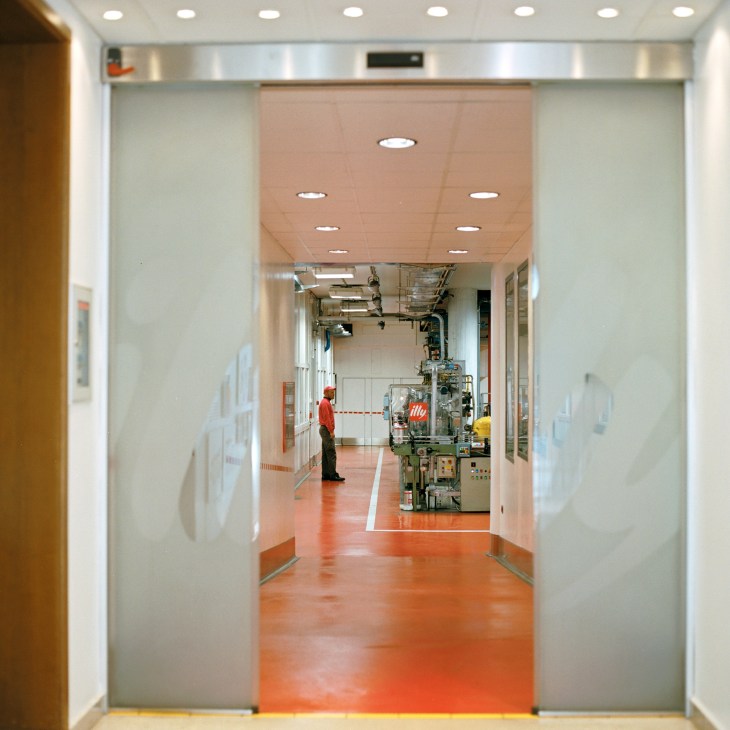 Modern entrance to the Illy coffee factory with glass doors opening onto an orange-floored production area with machinery and an employee in red.