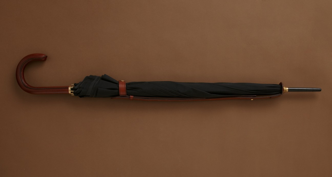 Rain Man umbrella with black canopy, red leather strap and curved wooden handle on tan background.