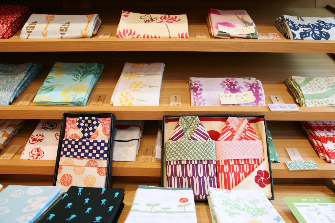 Wooden shelves displaying folded hand-dyed tenugui cloths and baby yukata in various colorful patterns at Kamawanu.