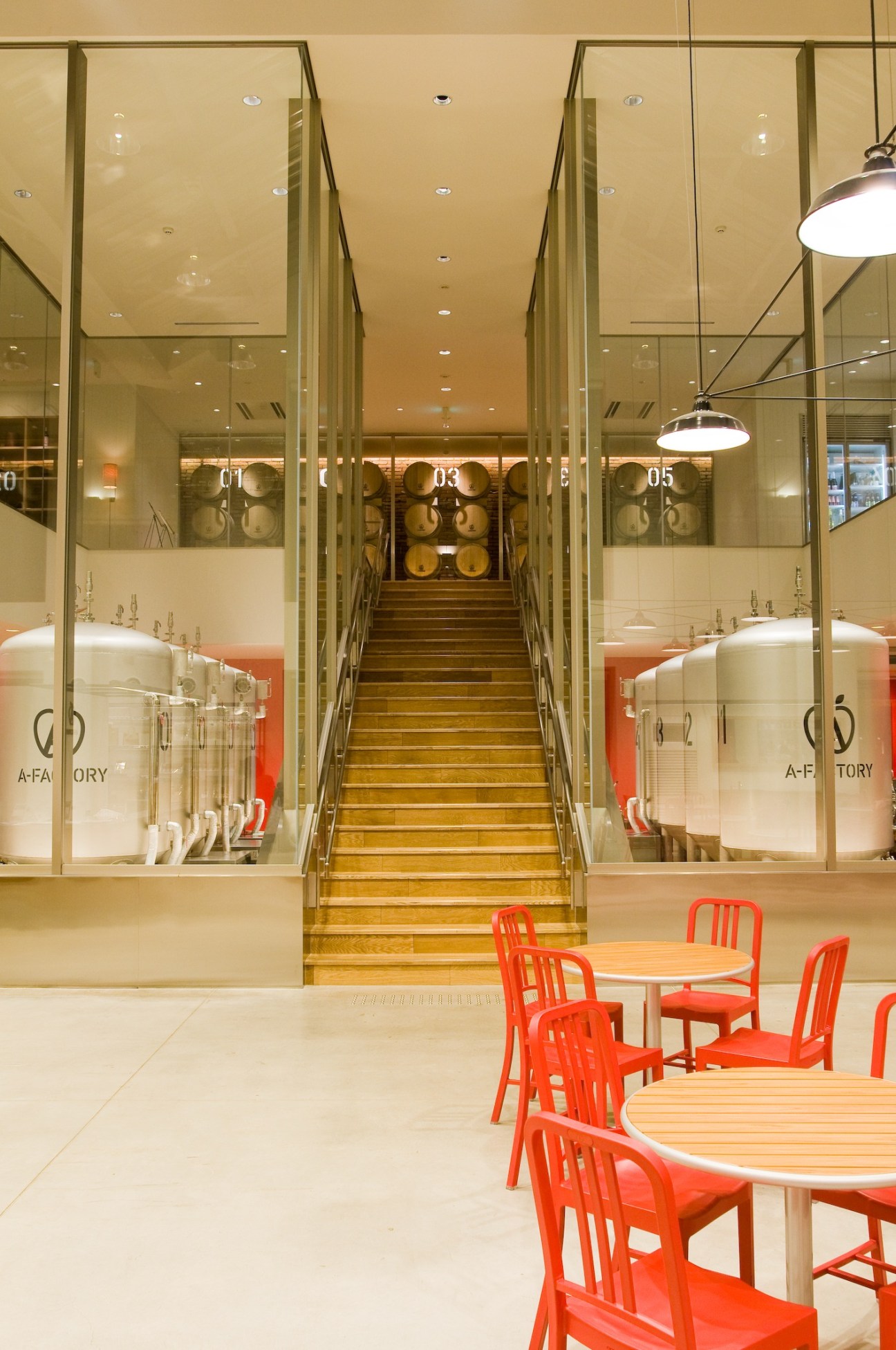 Interior stairway with wooden steps leading upward, flanked by glass-walled cider production tanks labeled A-Factory, with red chairs and tables in foreground.