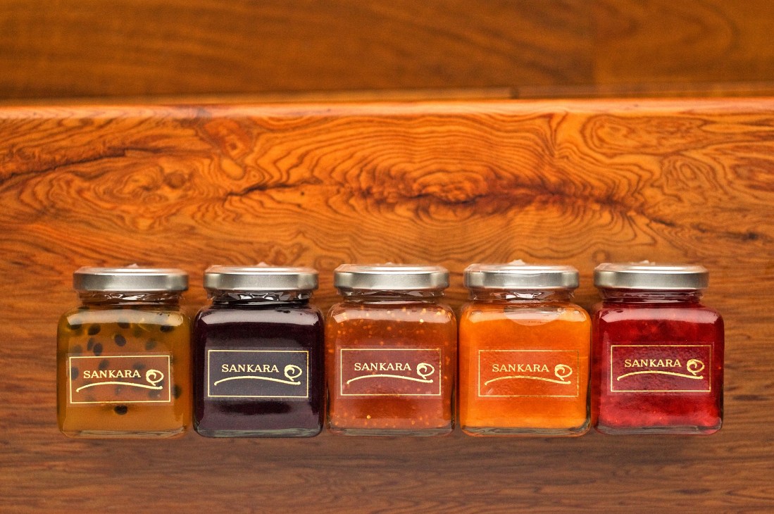Five glass jars of Sankara-branded organic jams in varying colors displayed on wooden surface.