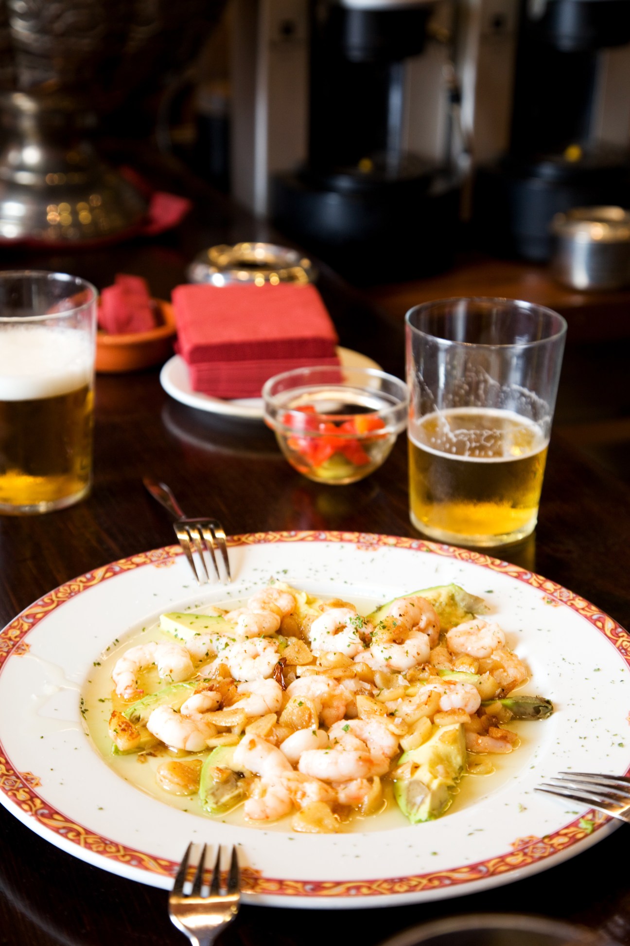 Plated seafood dish with shrimp and vegetables alongside glasses of beer and cocktails on a dark table.