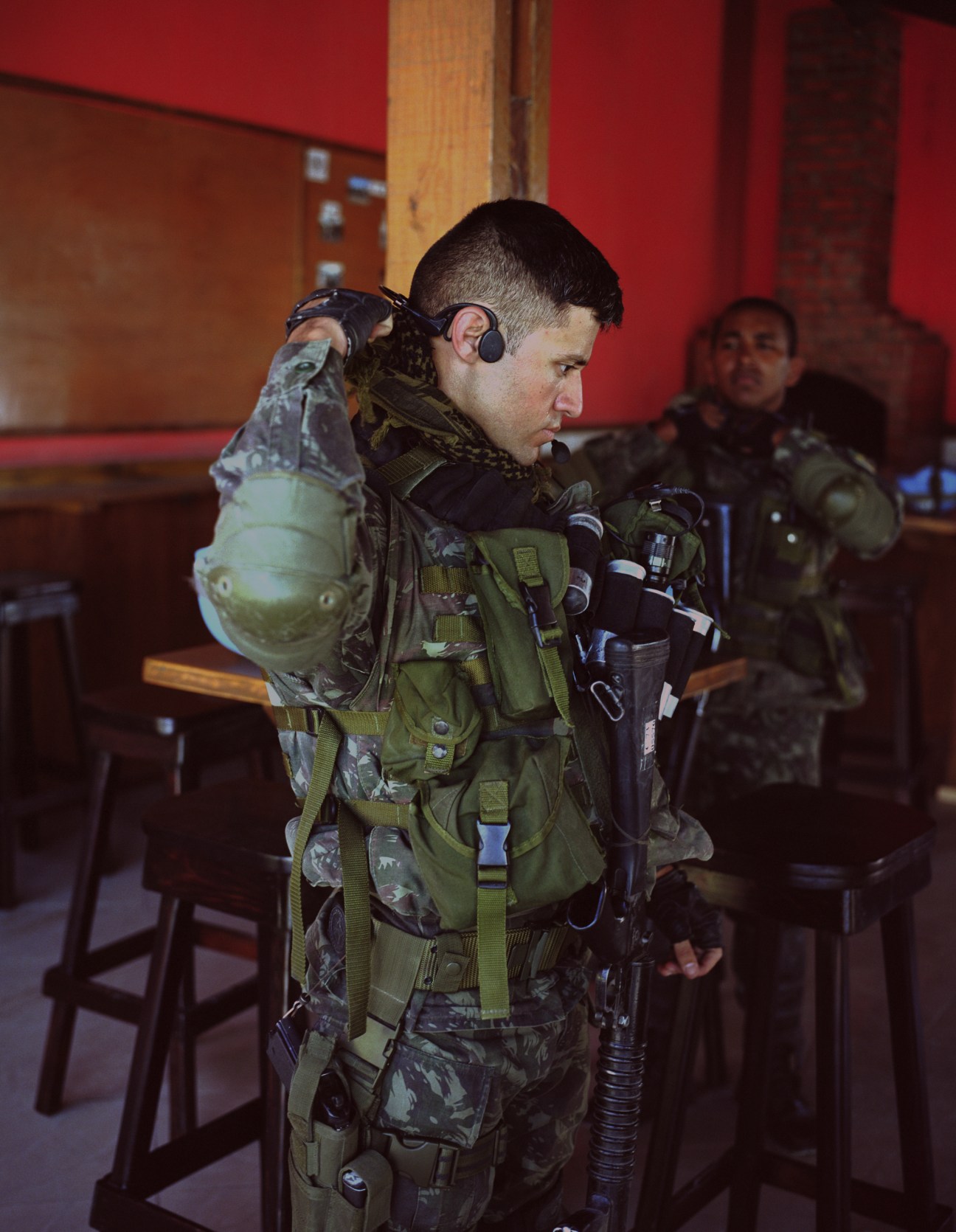 Brazilian soldier in full tactical gear preparing for patrol, adjusting helmet and equipment indoors.