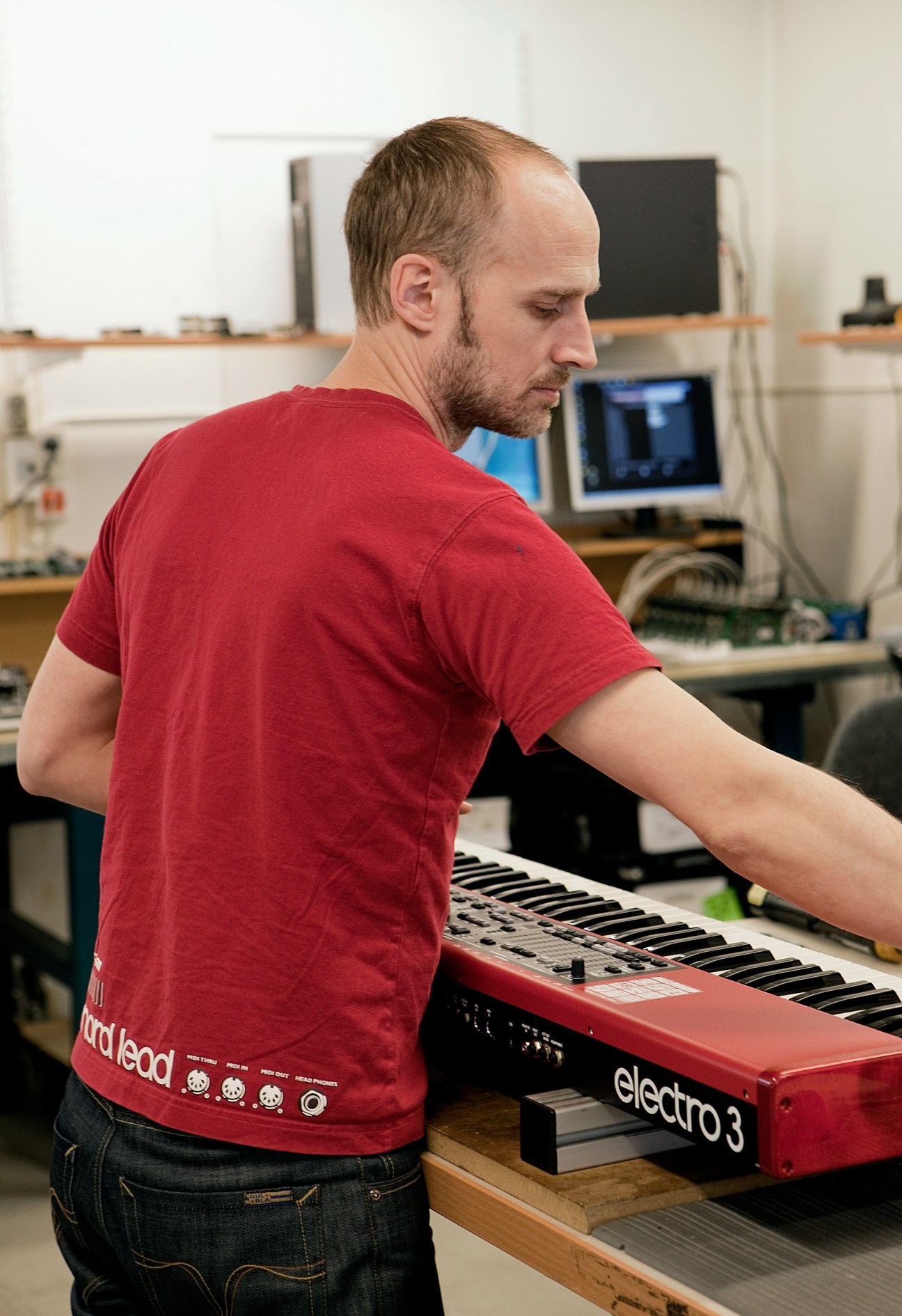 Man in red shirt playing a red Nord electro3 keyboard in a workshop with computers.