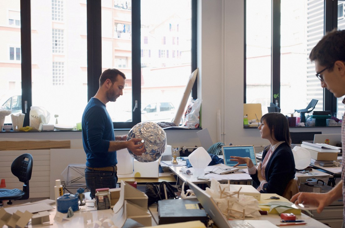 Professor Elric Petit evaluates a student's spherical design prototype in a bright workshop with city views.