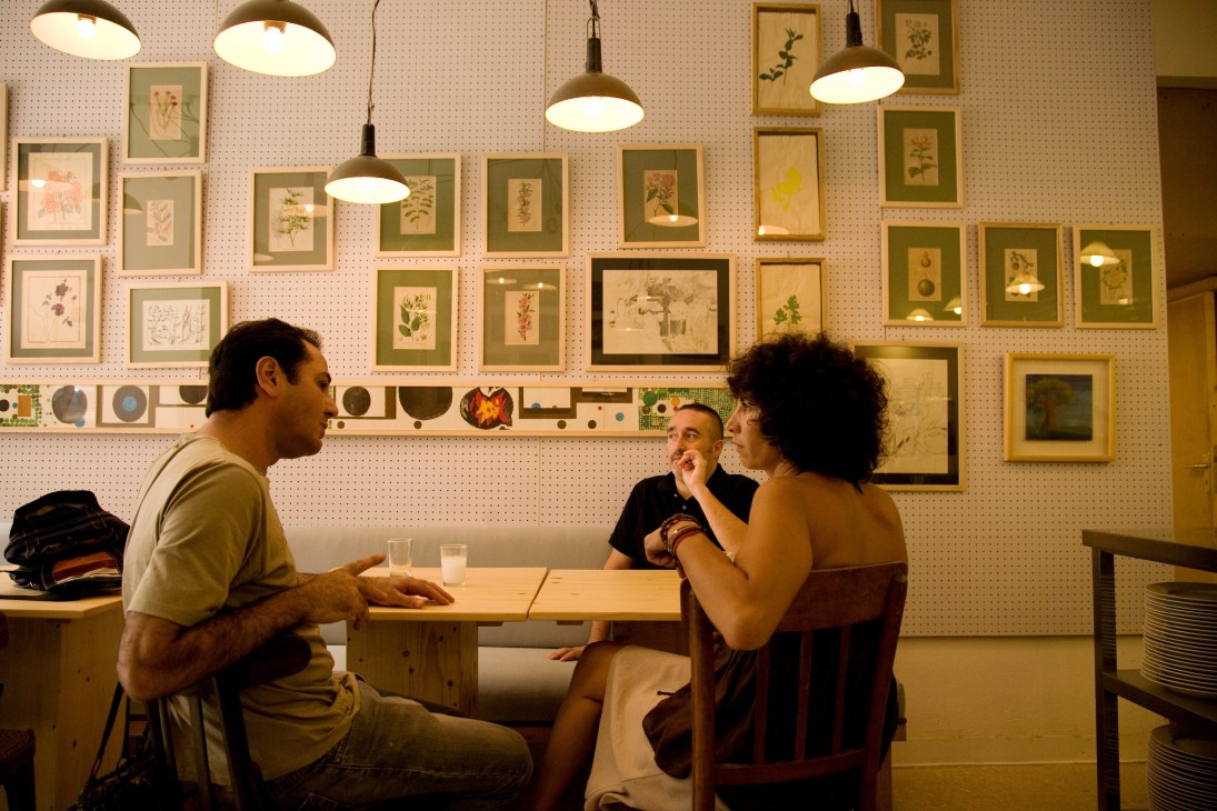 Three people seated at a wooden table in a minimalist restaurant with framed botanical prints on a pegboard wall and pendant lighting.