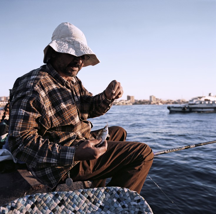 Fisherman holding a catch at the passenger terminal in Port Said, Egypt, with boats and waterfront buildings visible behind him.