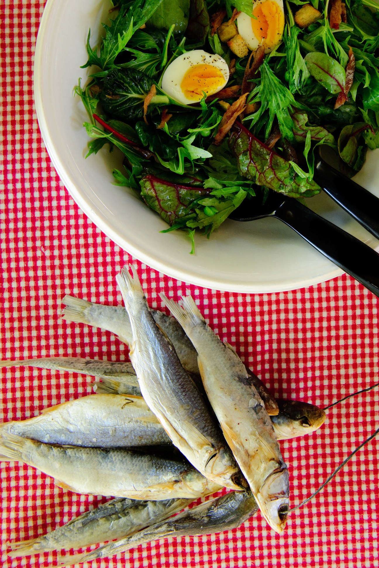 Salad with greens and soft-boiled egg beside dried mullet fish on red gingham tablecloth.