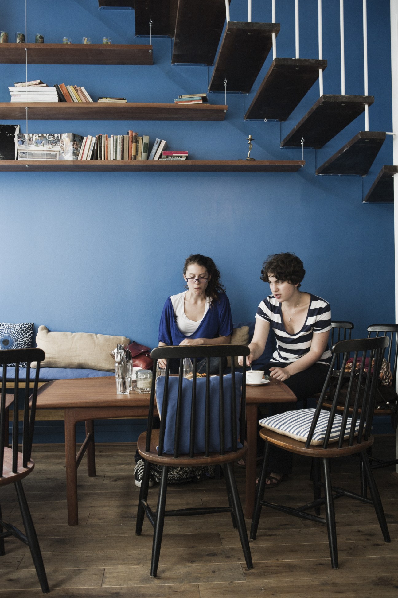 Two women sit at a wooden table in a bright blue café with floating shelves and black chairs.