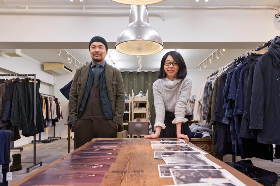 Two people stand in a modern clothing store with racks of dark garments and a large metallic pendant lamp.