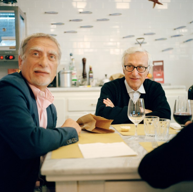 Two men seated at a table in a modern restaurant with white ceiling and bar counter behind them.