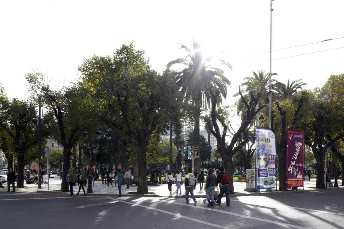 Pedestrians walk through Plaza Victoria in Valparaíso with tall trees, palm trees, and buildings in the background.