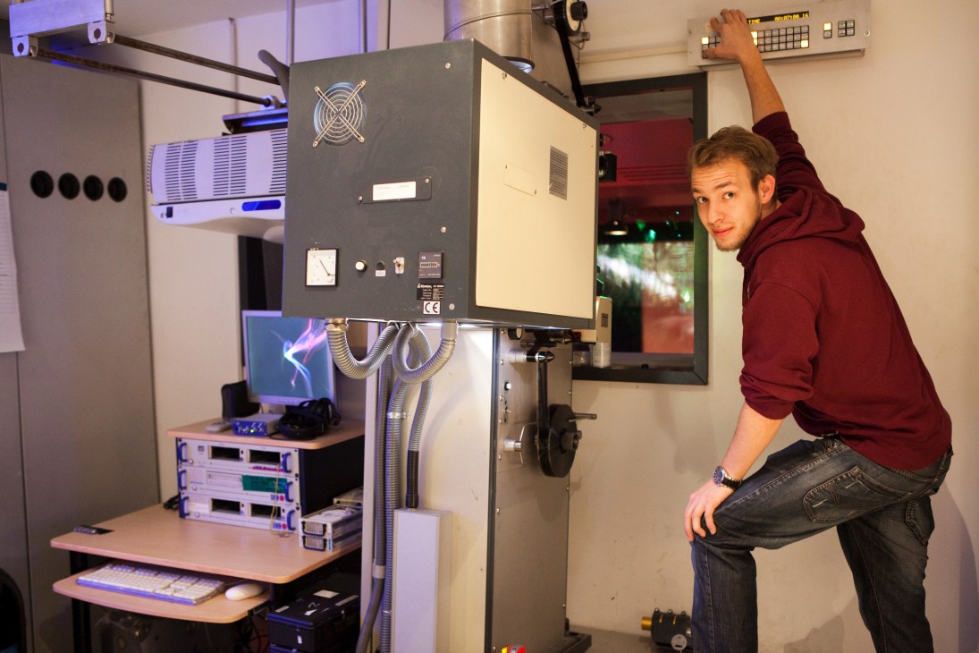 Man in maroon hoodie gestures toward screening equipment in a professional dubbing studio with monitors and audio gear.