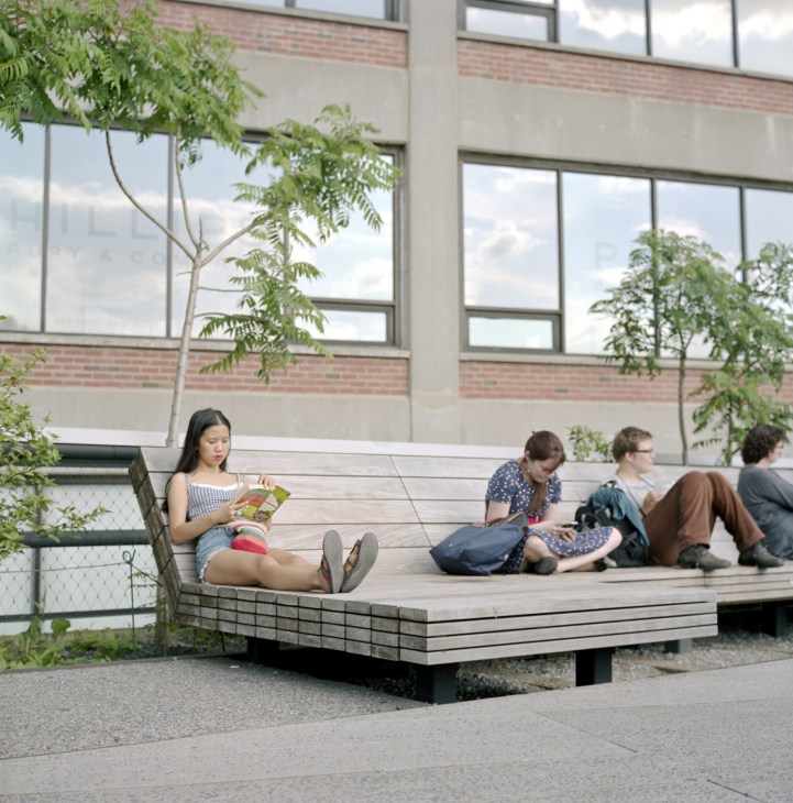 Young people sitting and relaxing on a wooden platform bench with greenery and a brick building in the background.