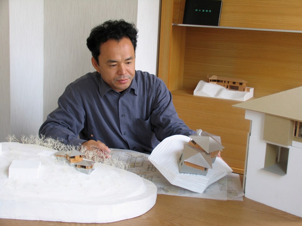 Japanese architect Yasushi Horibe examines architectural models at a wooden desk.