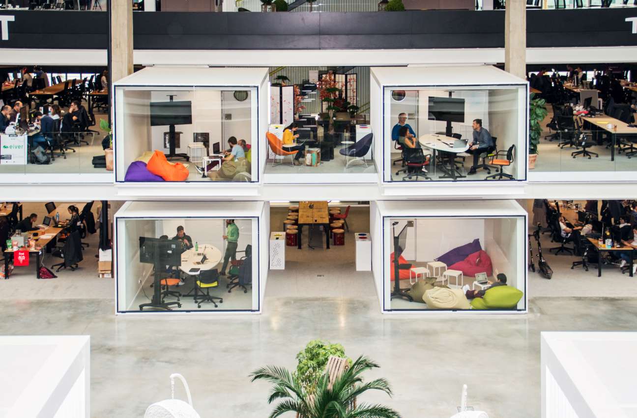 Multiple glass-walled private office pods in a modern co-working space with workers and colorful furniture visible inside.