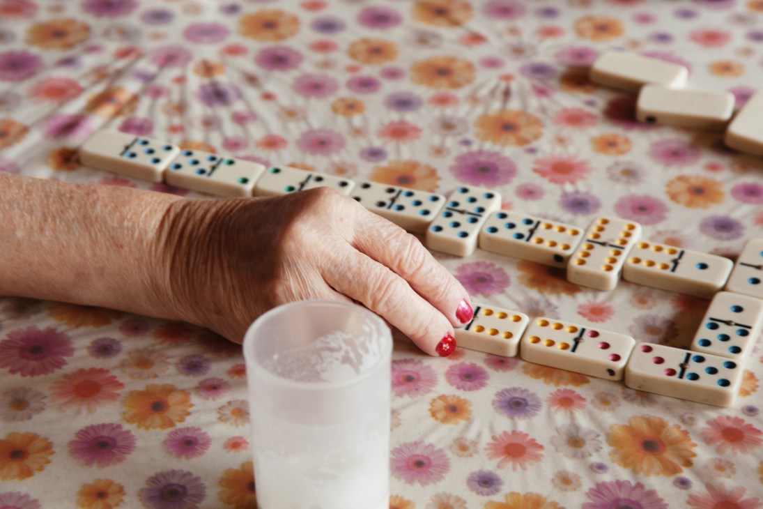 An elderly person's hand with pink nail polish playing dominoes on a floral patterned surface.