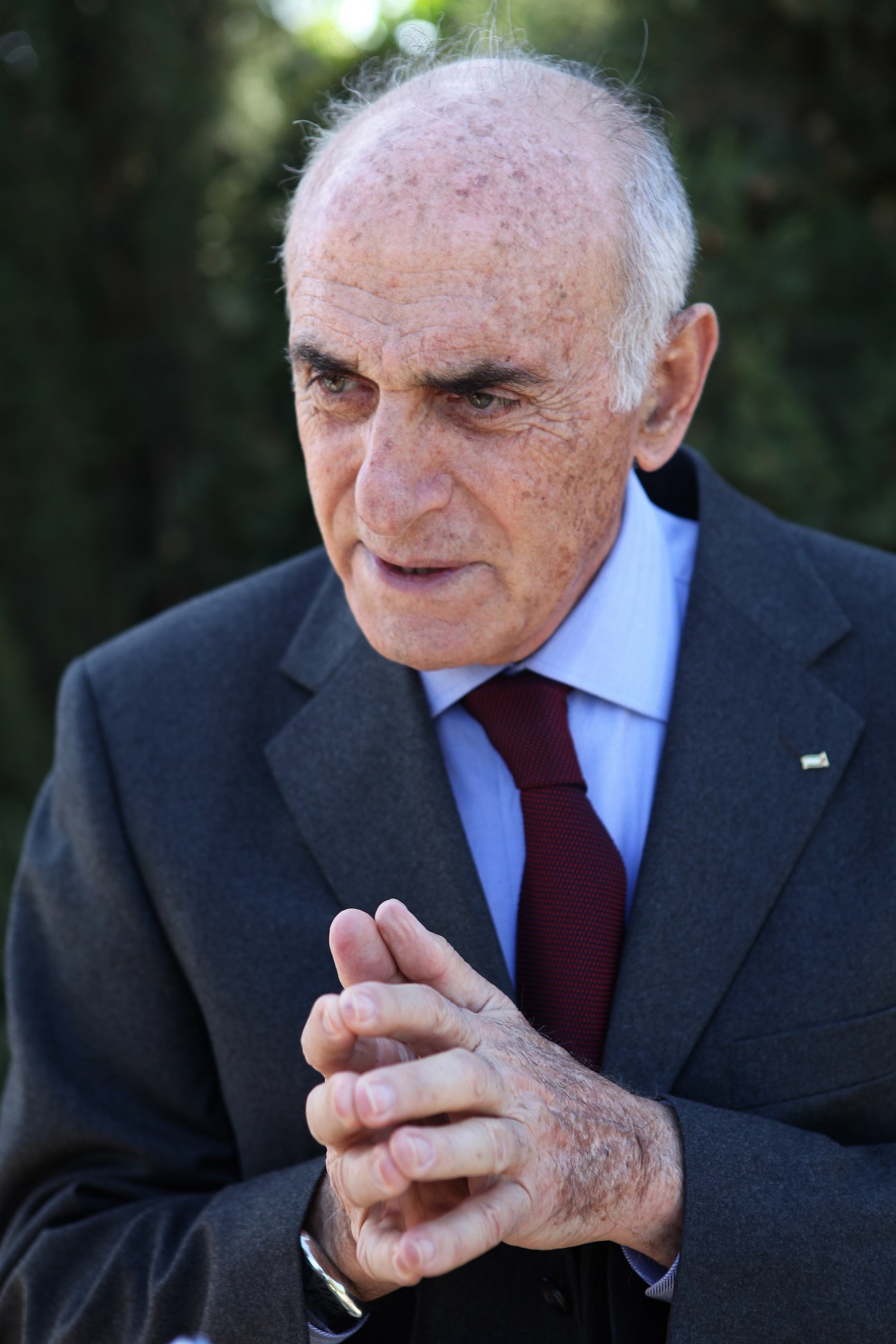 Munib al-Masri, an elderly man in a dark suit and burgundy tie, gestures while speaking outdoors.