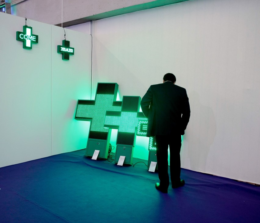 Man in dark suit viewing illuminated green pharmacy cross neon signs at trade fair booth.