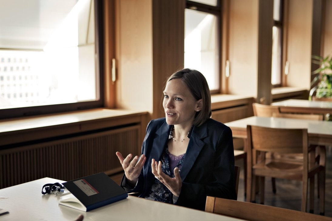 Woman in navy blazer gestures while speaking at a desk in a bright office with wooden window frames.