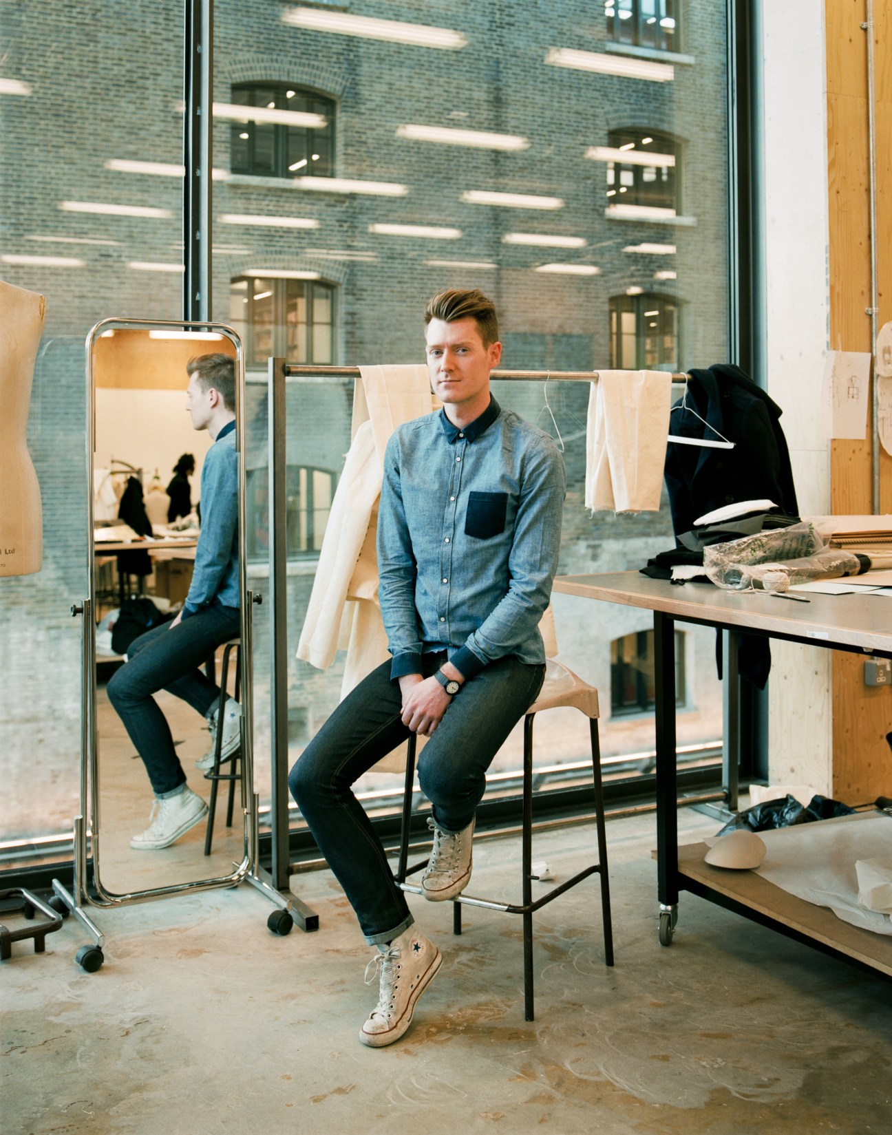 Male student in denim shirt sitting in menswear studio with fabric swatches and mirror.