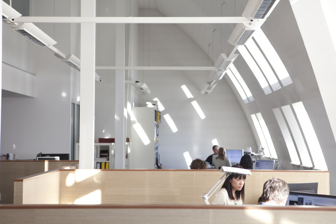 Bright open-plan office with skylights, wooden desks, and employees working in a minimalist space.