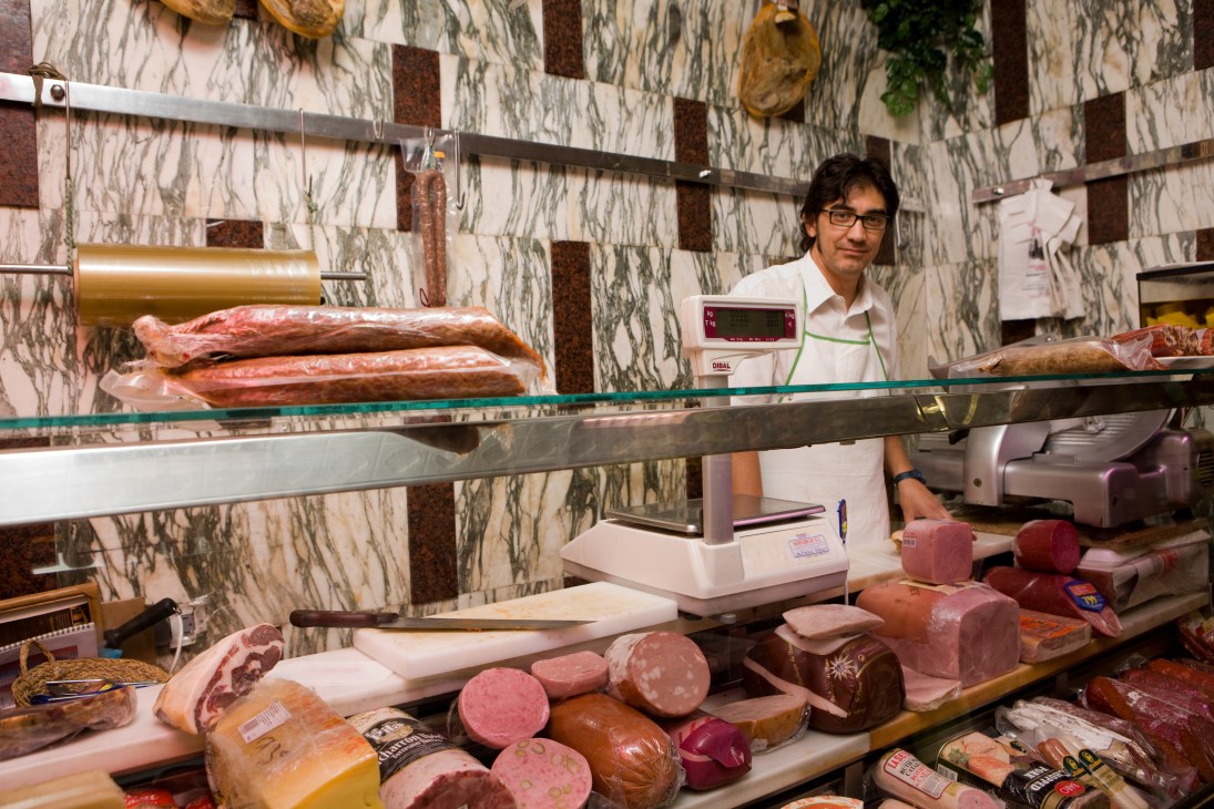 Carlos, manager of Guticar Alimentación, stands behind a glass counter displaying cold cuts, meats and sausages in a Madrid delicatessen.