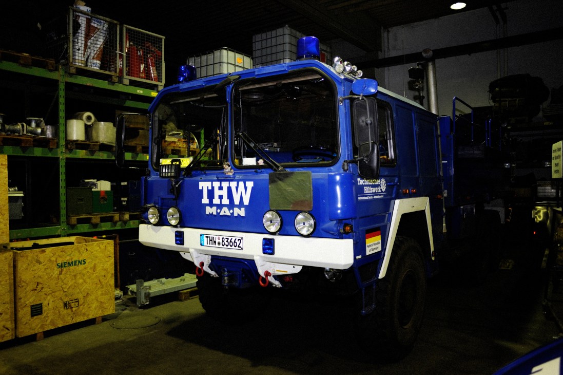Blue THW MAN truck parked in a warehouse garage with emergency equipment and supplies visible.