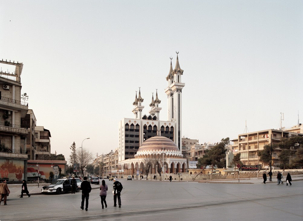 Al Rahman mosque with twin minarets and dome in Aleppo's main square, surrounded by modern buildings and pedestrians.