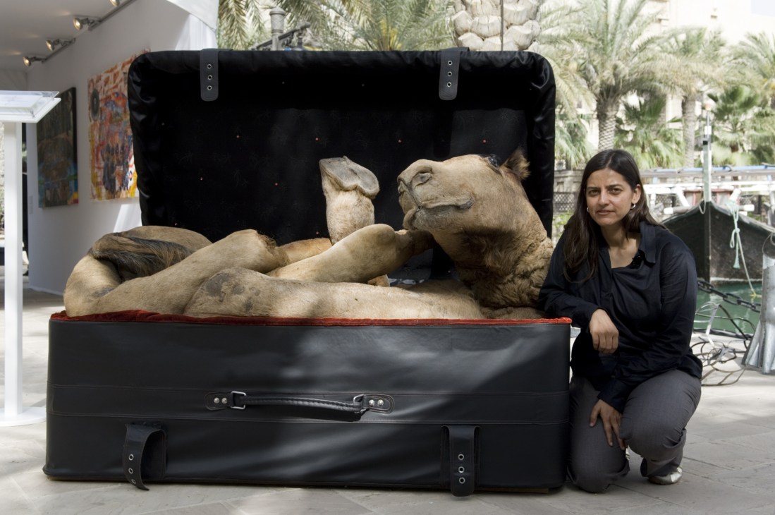 Artist Huma Mulji poses beside her taxidermied camel sculpture packed in a black suitcase.