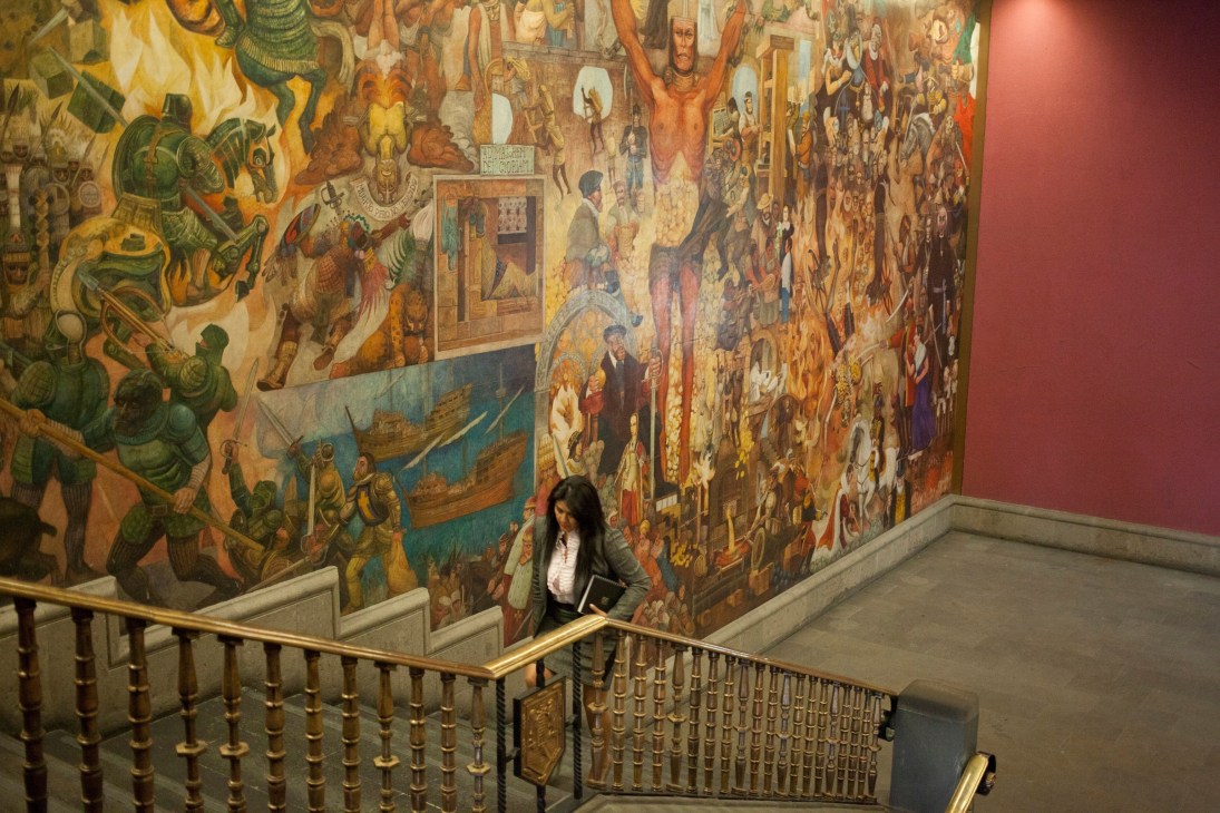 Woman descending staircase in National Palace with large colorful Mexican murals covering the walls