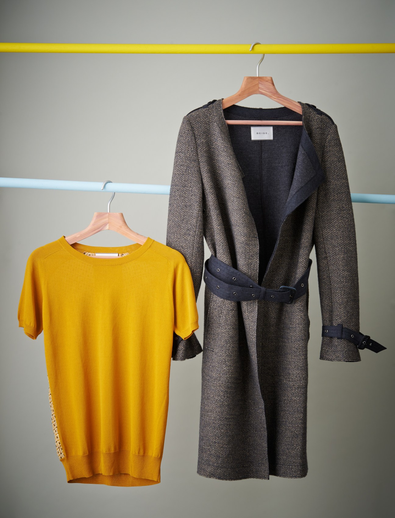 Yellow t-shirt and charcoal wool coat with black belt displayed on wooden hangers against a gray wall with colored pipes.