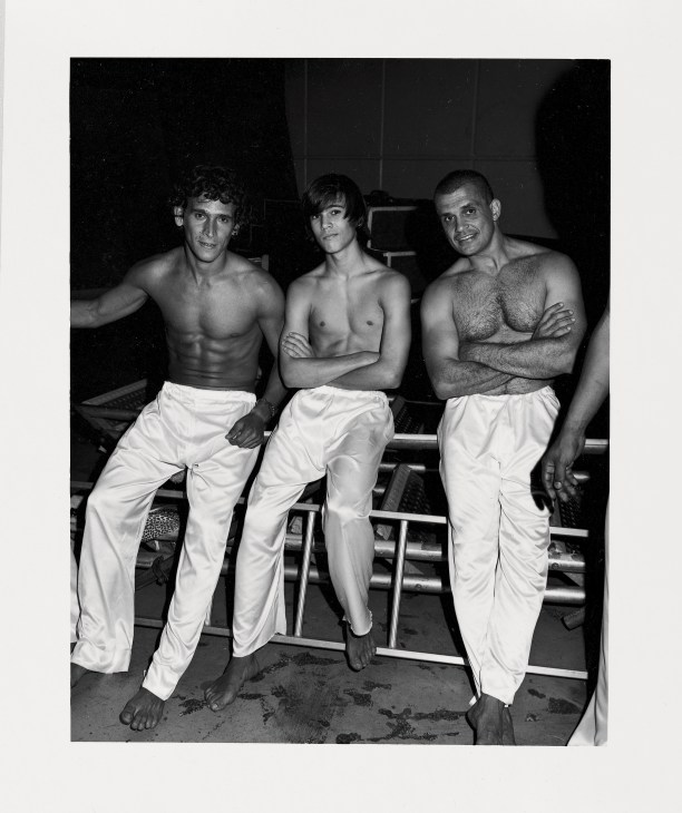 Three shirtless Brazilian capoeiristas in white pants sit together at the opening ceremony.