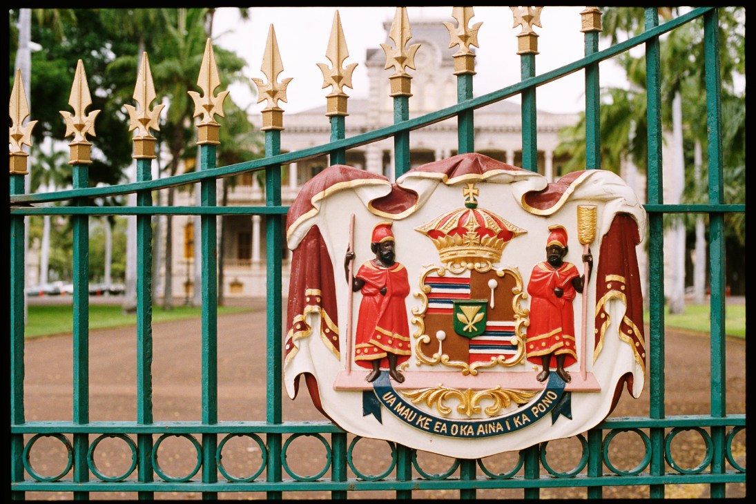 The Hawaiian coat of arms mounted on an ornate green and gold fence with palace gates behind.