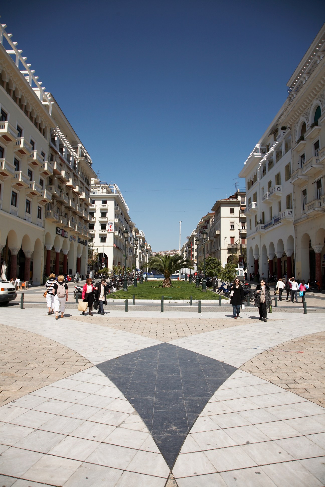 Aristotelous Square in Thessaloniki with neoclassical buildings, tree-lined promenade and pedestrians under clear blue sky.