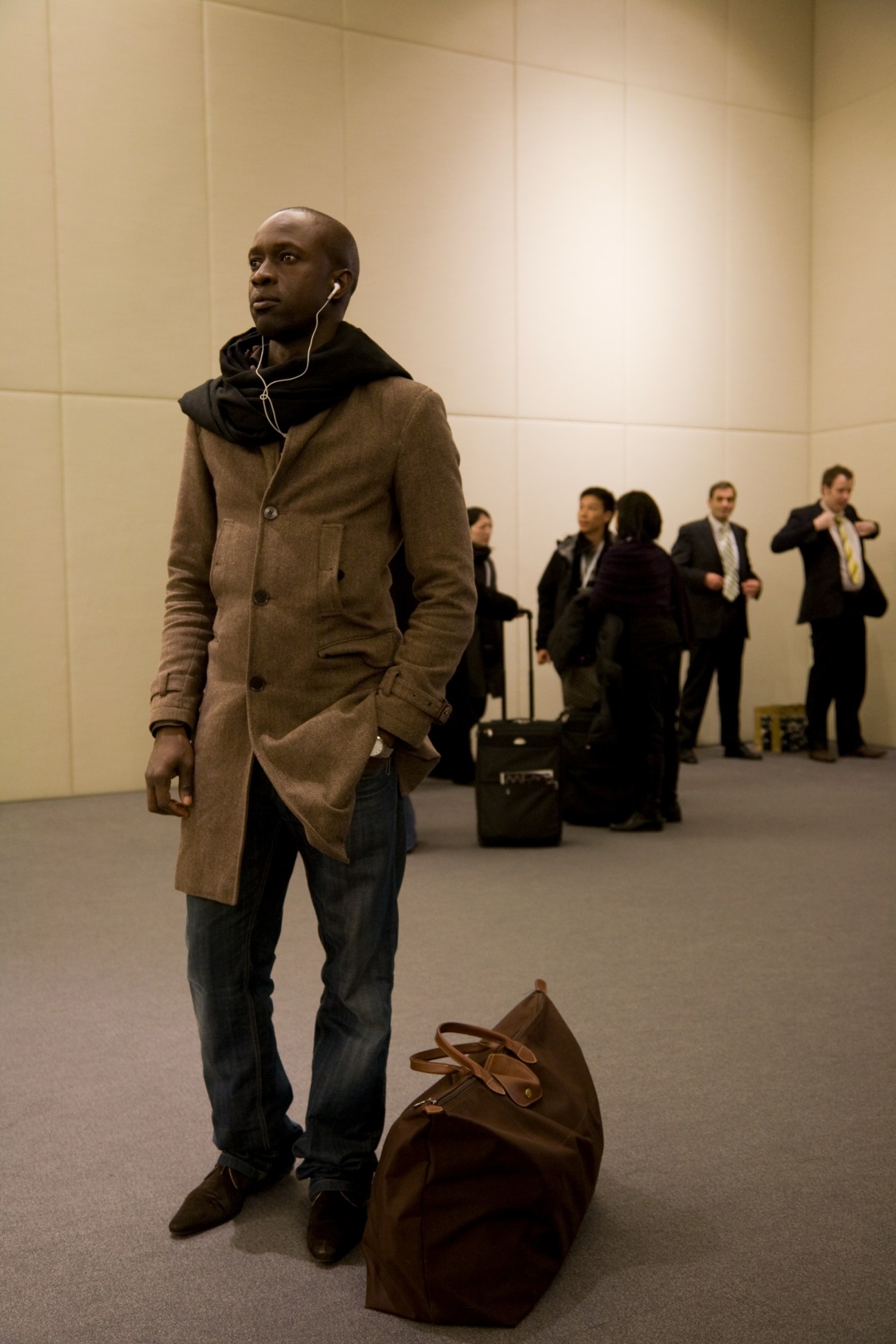 Well-dressed man in tan coat and earbuds standing in gallery space with other attendees and luggage.