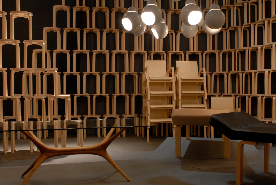 Artek exhibition stand featuring wooden chairs stacked on wall, pendant lights, and modern furniture displays