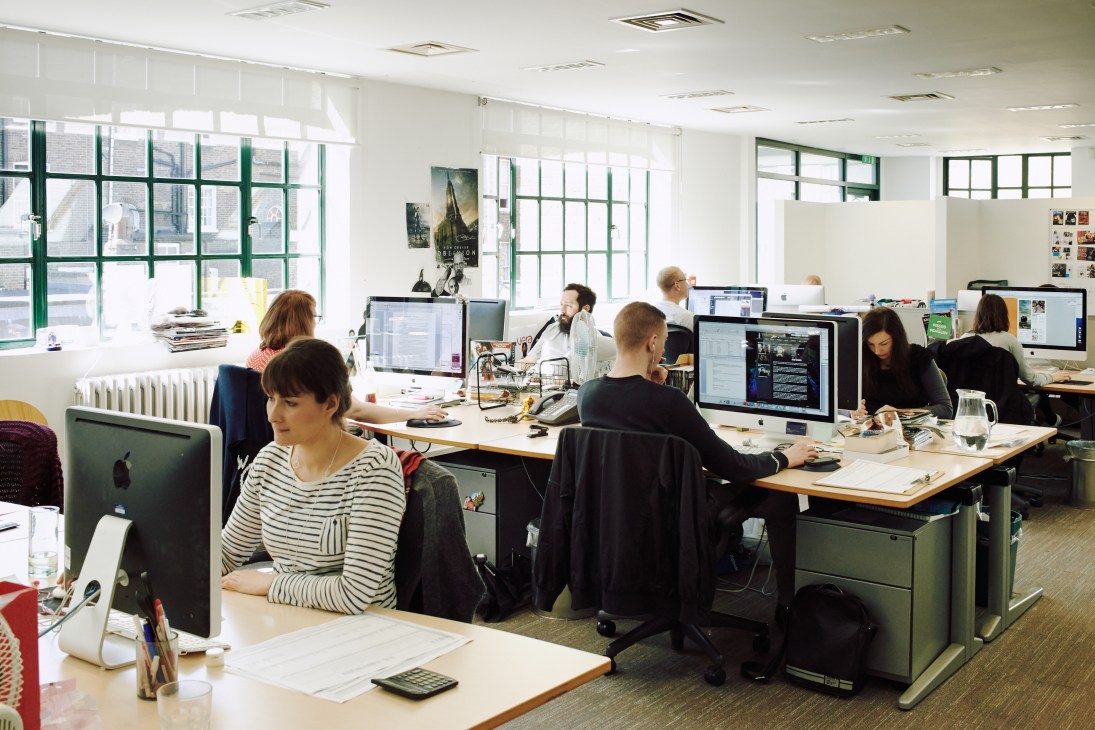 Open-plan office with staff working at desks with computers, green-framed windows, and bright natural light at Spafax London.