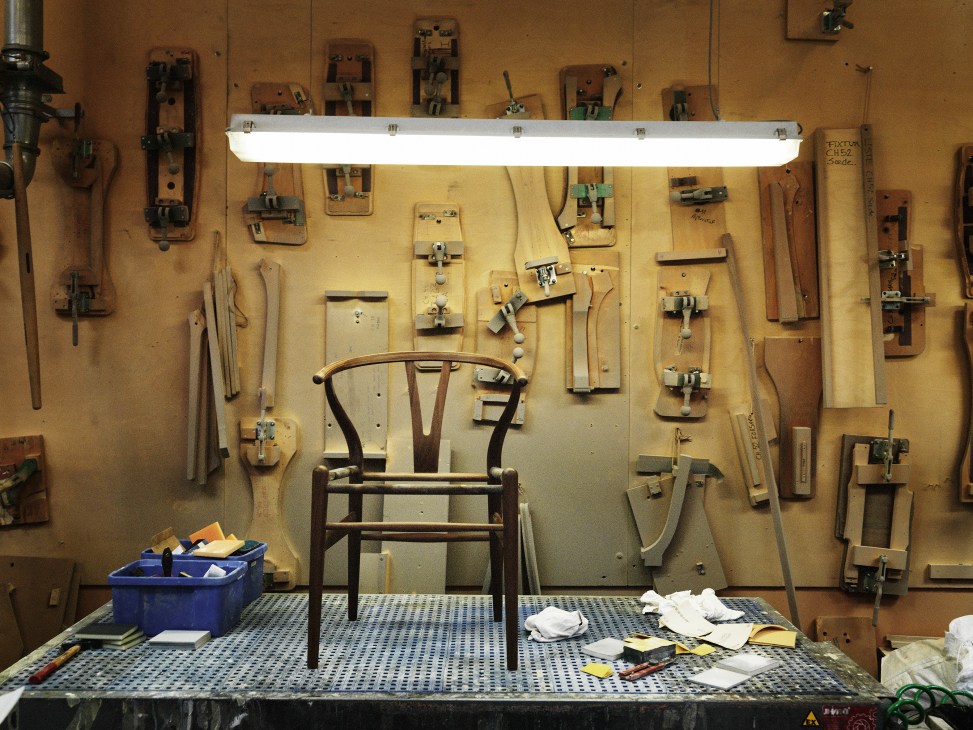 A wooden Wishbone chair displayed on a workbench surrounded by woodworking tools and fixtures mounted on a yellow wall.