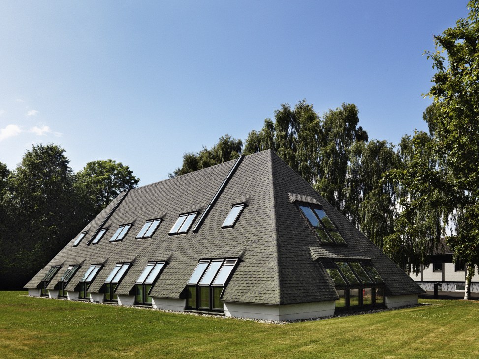 Modern angular building with dark slate roof and large windows set in a green landscape.