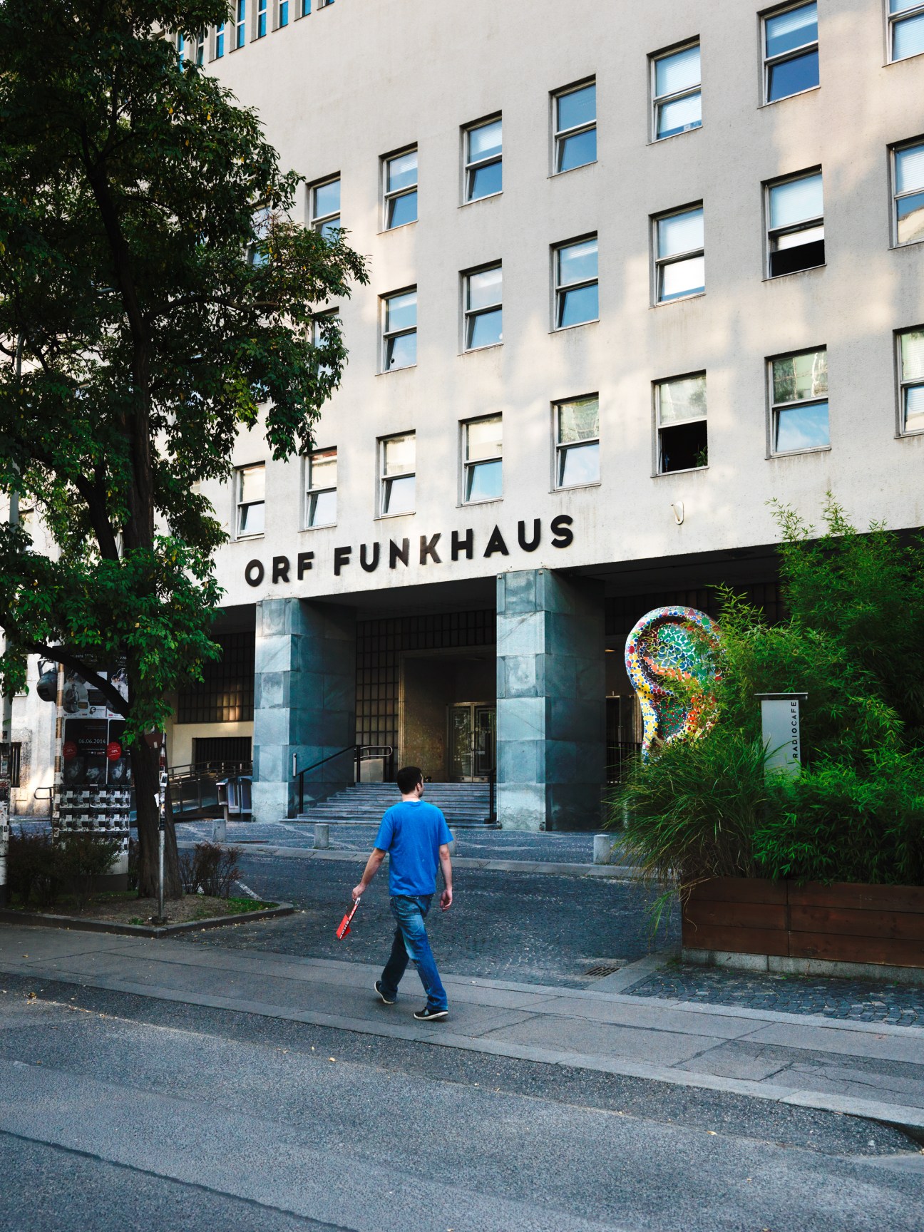 Person in blue shirt walking past ORF Funkhaus building in Vienna with colorful head sculpture visible.