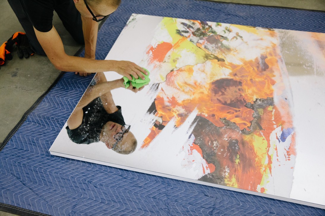 A worker's hands clean a colorful abstract artwork with orange, yellow and red tones on a blue patterned floor.