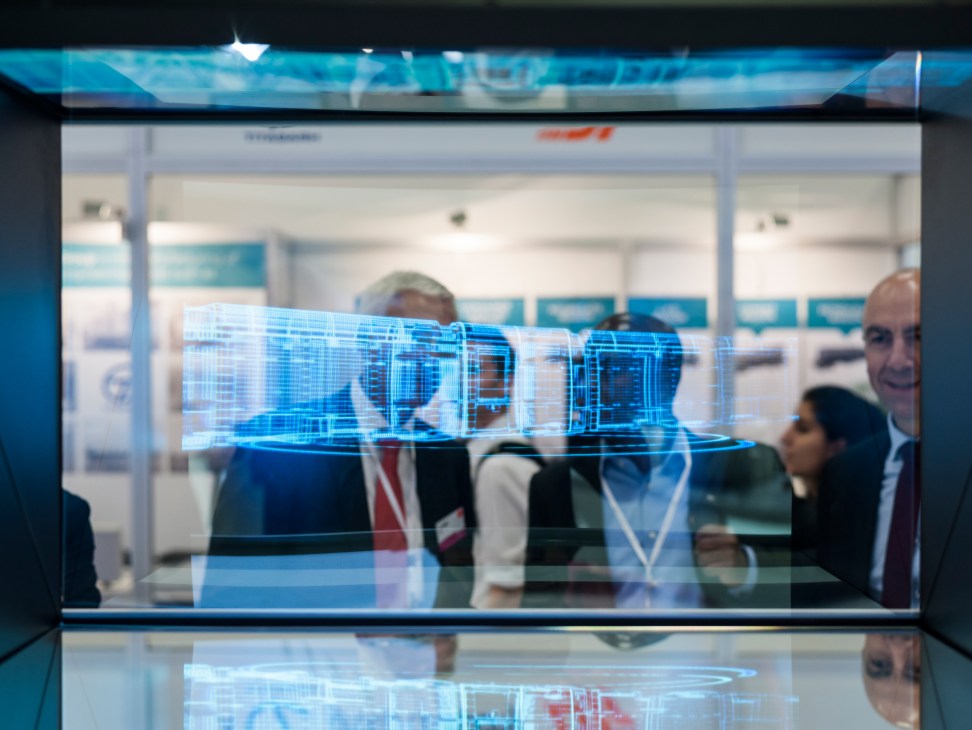 A holographic display of a blue train model at the Bombardier booth, with visitors visible in the background.