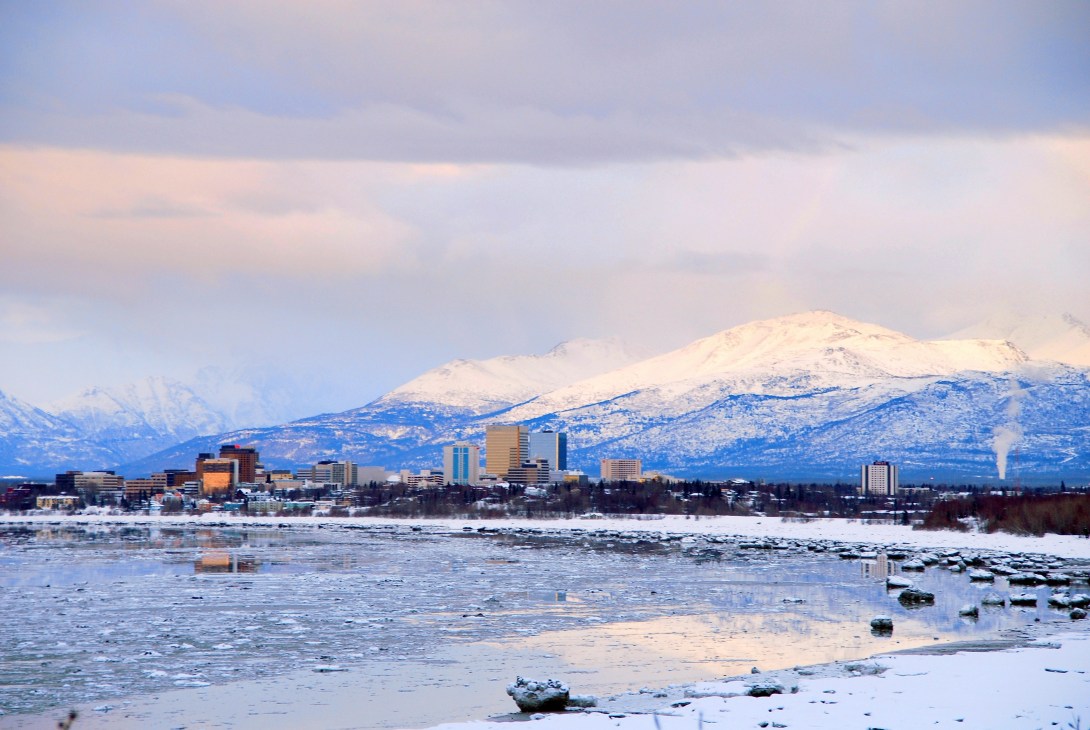 Anchorage skyline with snow-capped mountains reflected in frozen water below.