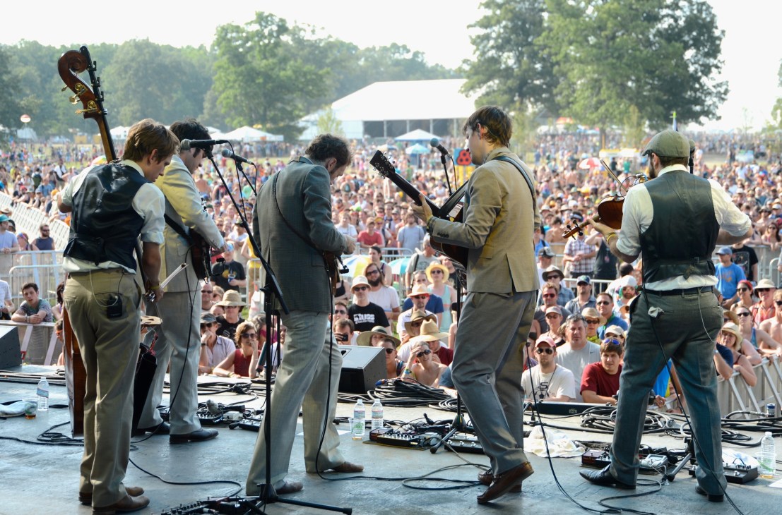 Band performs on stage at Bonnaroo festival with large crowd and white tent in background.