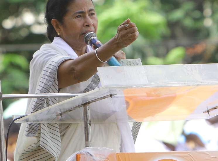 Mamata Banerjee speaking at a podium with microphone, gesturing while addressing an audience outdoors.