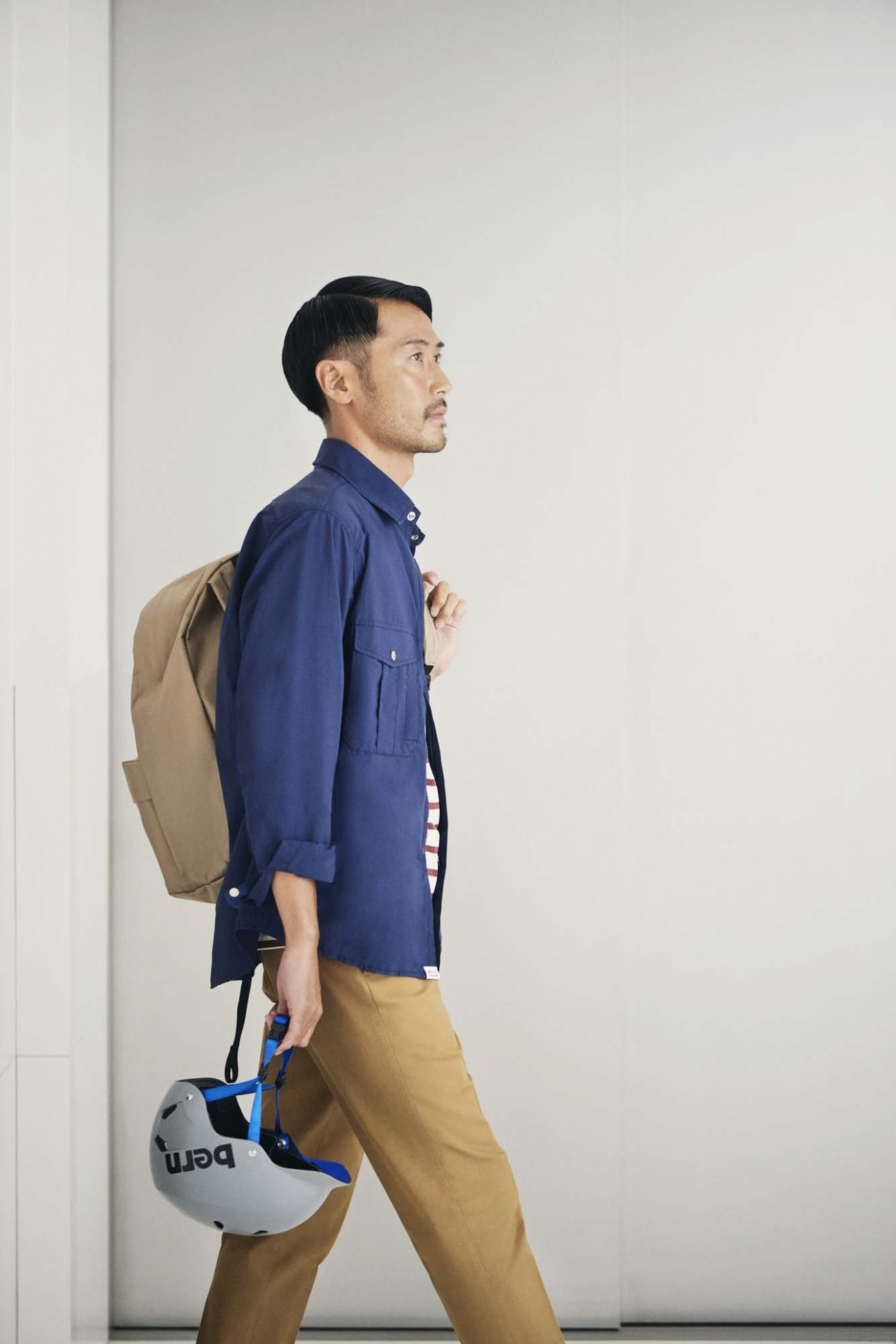 Man wearing navy shirt jacket, checkered shirt, tan trousers, holding a backpack and safety helmet.
