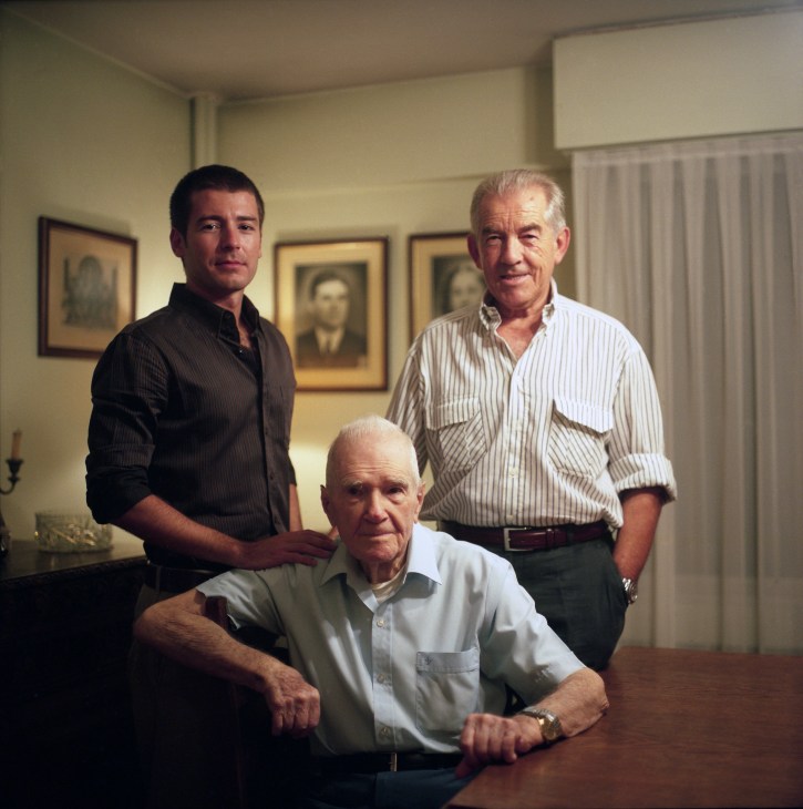 Three men of different generations pose indoors, with framed portraits hanging on the yellow wall behind them.